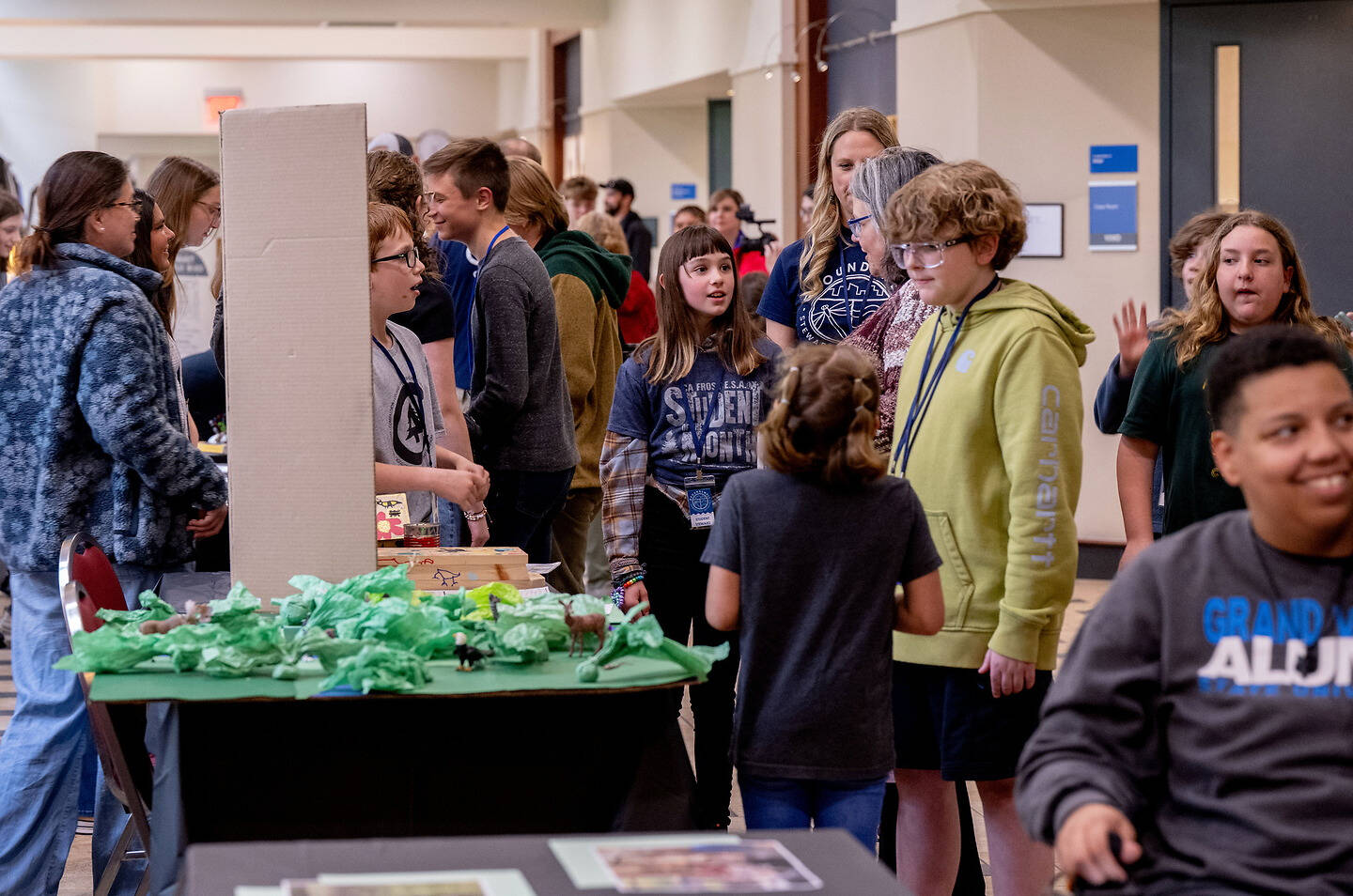 A group of young people gather around a table displaying a handmade diorama, engaging with each other and observing the exhibit.